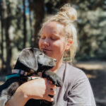A woman of Caucasian descent smiles, holding her black Labrador Retriever puppy while hiking outside on a Summer day.